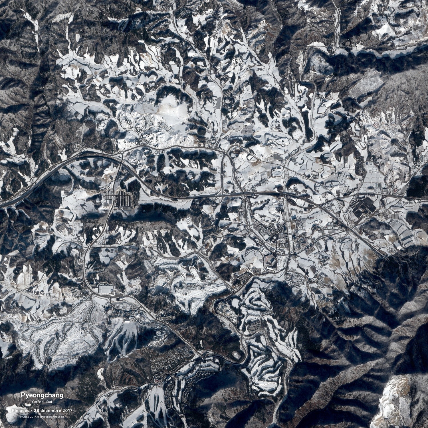 Une ville enneigée vue depuis l'espace. Elle semble située entre les plis d'une montagne. Une autoroute coupe la photo en deux.