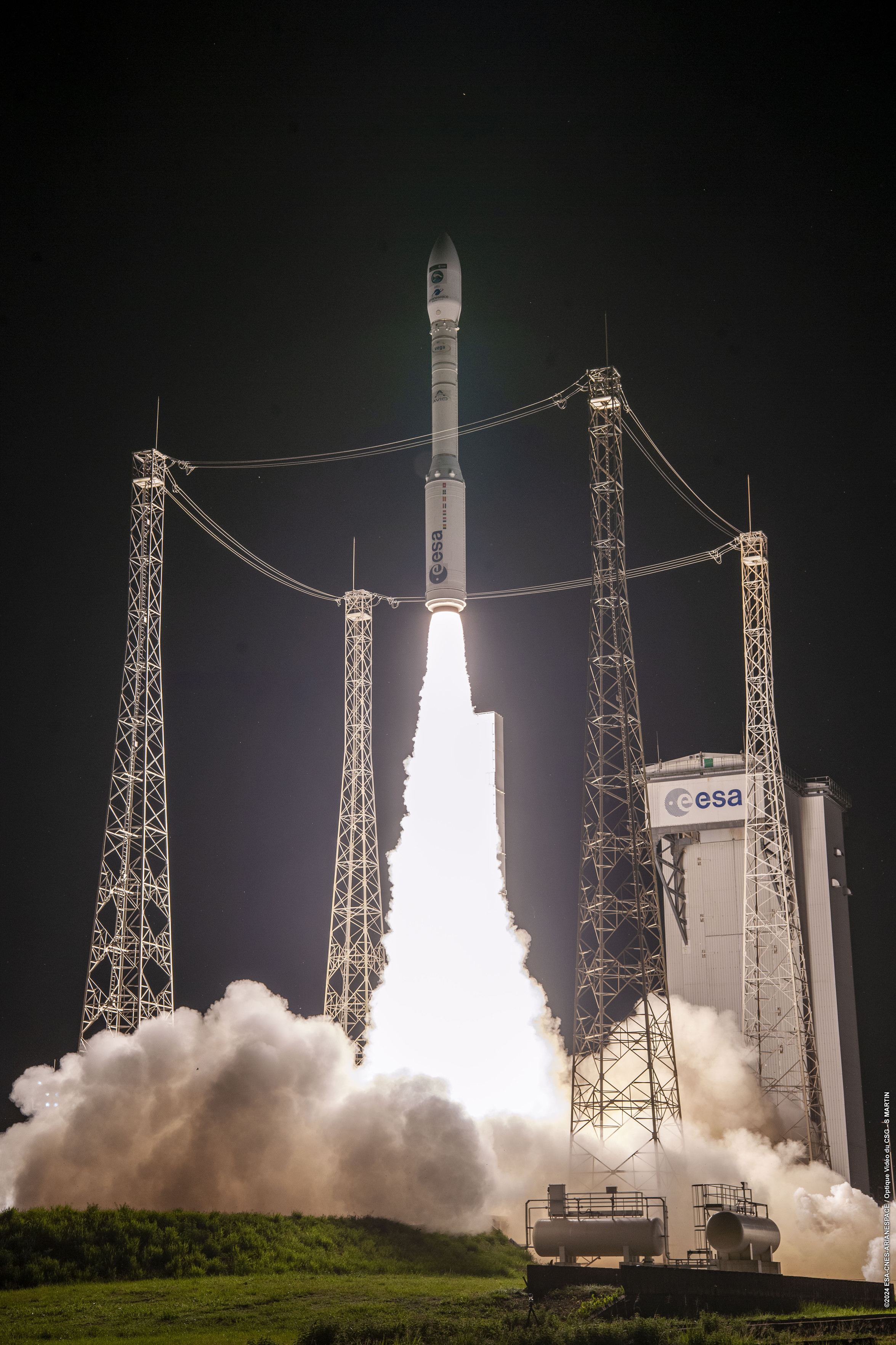 Vega au décollage, au moment où elle va dépasser les paratonnerres. La flamme très lumineuse illumine le pas de tir, le portique mobile à l'arrière plan et l'herbe verte au premier plan. Le ciel est très sombre.